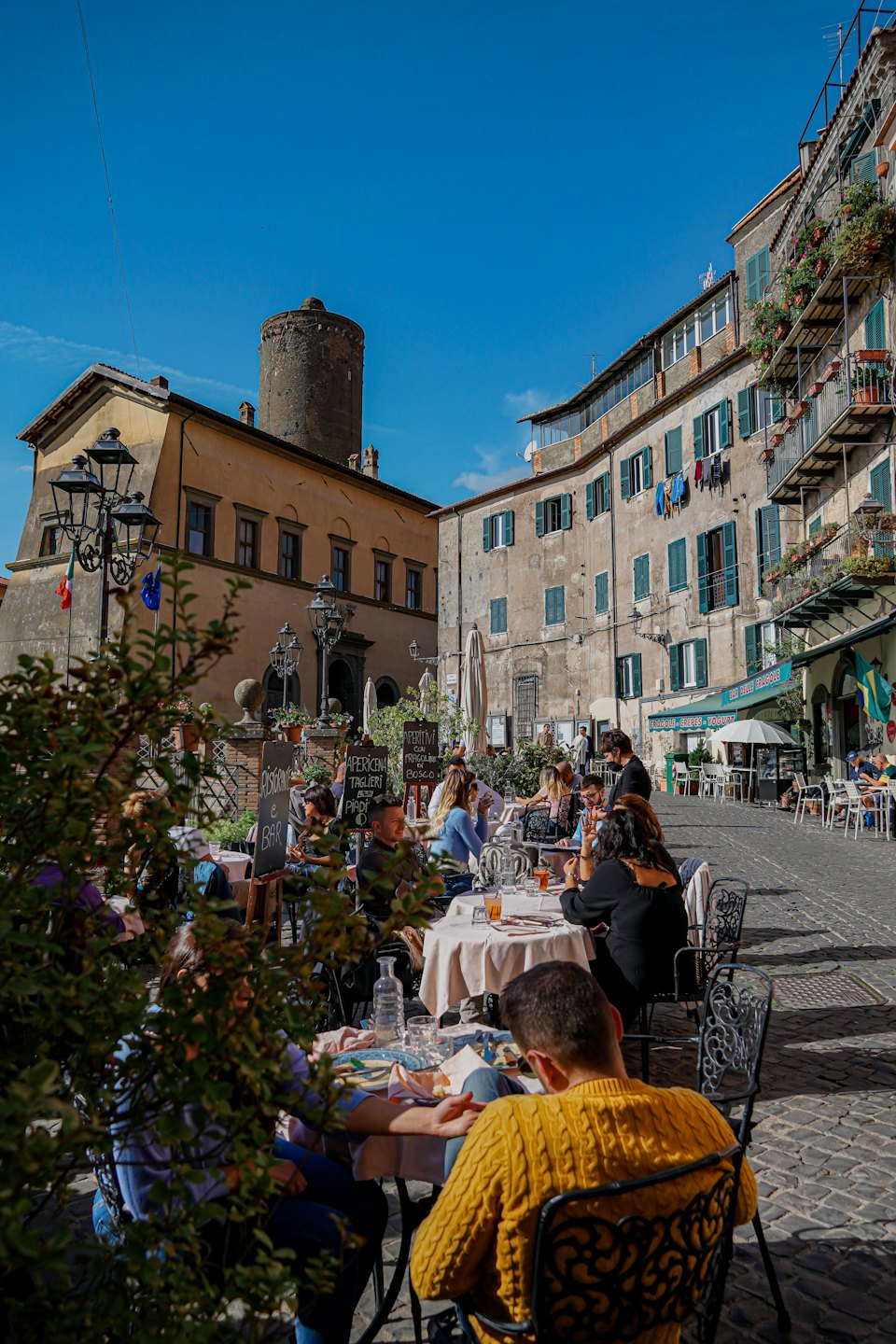 Persone cenano a tavoli all'aperto in una storica piazza acciottolata.
